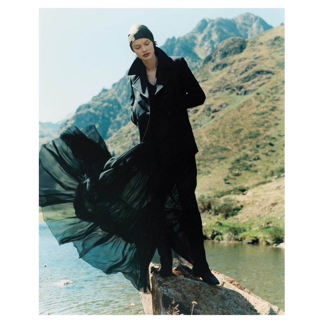 Fashion shot of a woman in a black coat and dress standing on a rock near a river with mountains in the background, outdoors.