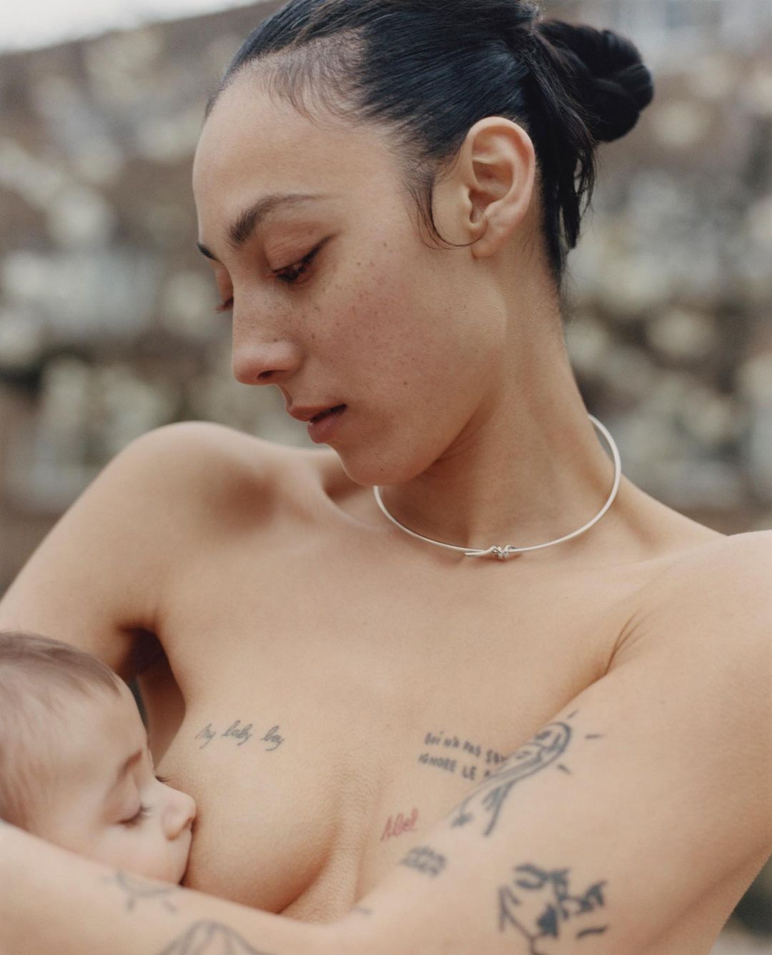Adult female breastfeeding a baby in a close-up studio shot, tattoos visible on the woman's body and a stone wall background.