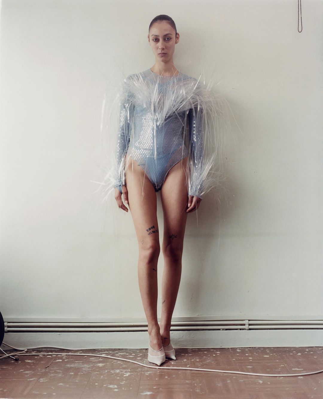 A slender adult female wearing a blue sequined bodysuit and a clear plastic garment stands indoors in a full shot against a wall.