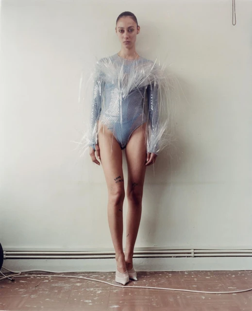 A slender adult female wearing a blue sequined bodysuit and a clear plastic garment stands indoors in a full shot against a wall.