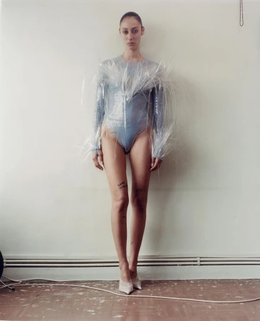 A slender adult female wearing a blue sequined bodysuit and a clear plastic garment stands indoors in a full shot against a wall.