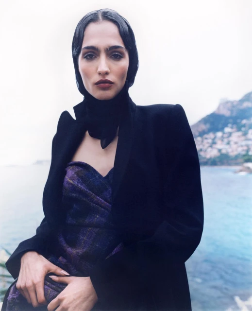Adult female with head covering and dark blazer poses outdoors against a backdrop of water and mountains on an overcast day.