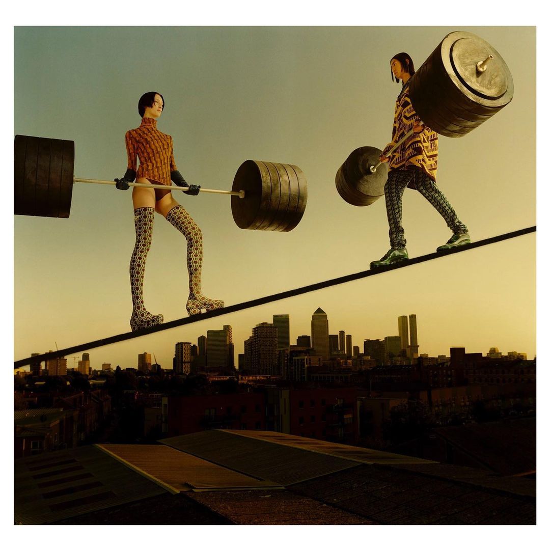 Two figures balance on a tightrope between buildings in a cityscape, holding heavy barbells. The artistic image highlights balance and strength.