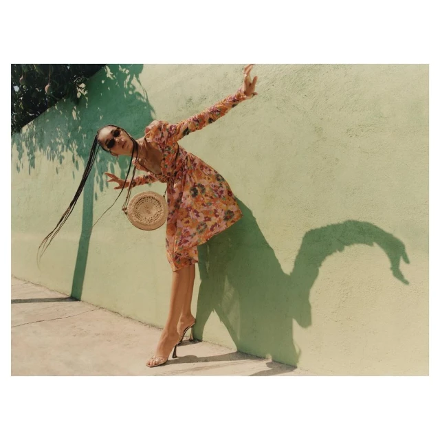 A woman in a floral print dress poses playfully against a light green wall on a sunny day, casting a large shadow, accessorized with a round bag.