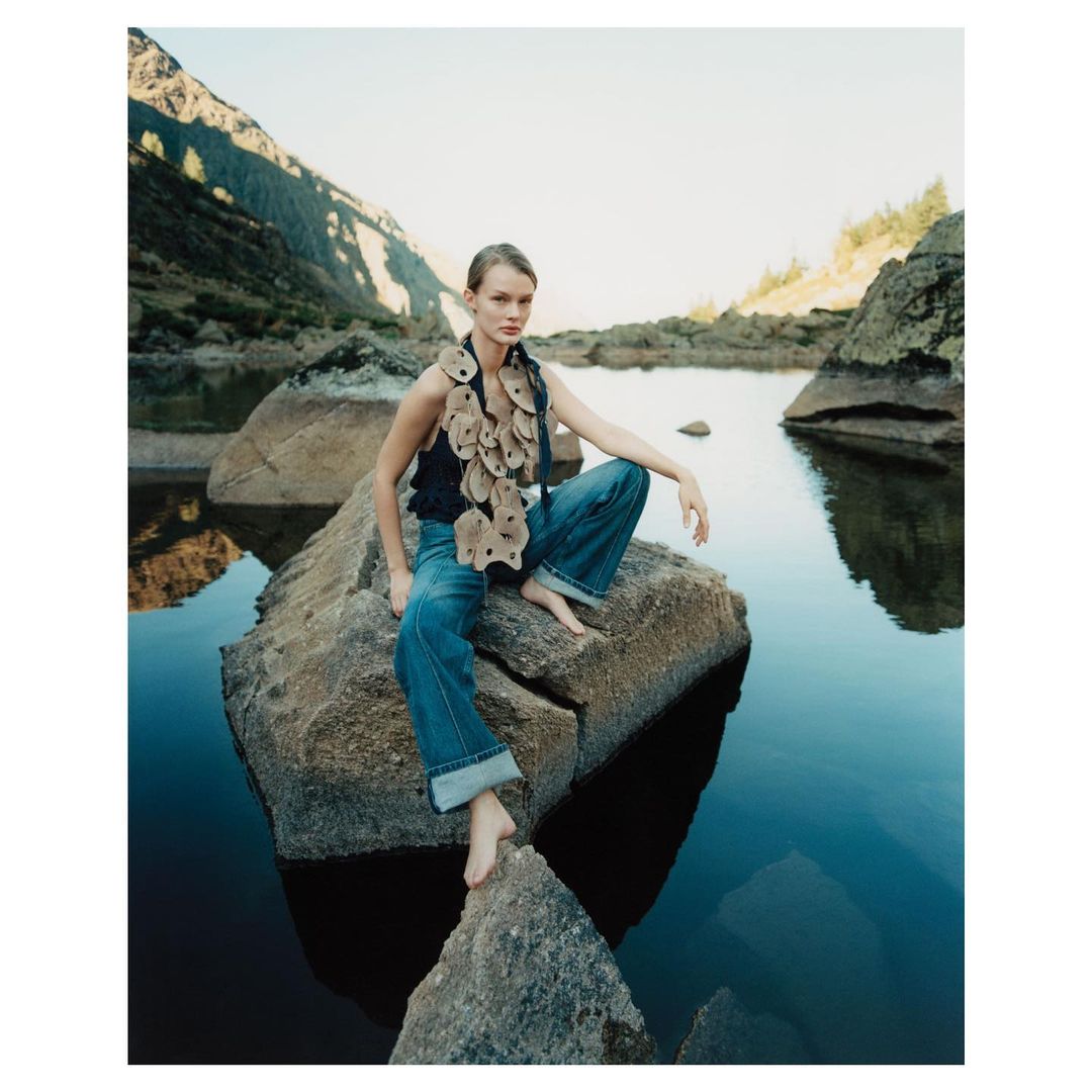 Woman in fashion denim and artisanal top poses on a rock with lake and mountain vista in the background in a lifestyle fashion shoot.