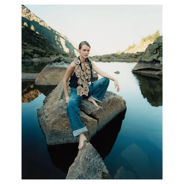 Woman in fashion denim and artisanal top poses on a rock with lake and mountain vista in the background in a lifestyle fashion shoot.