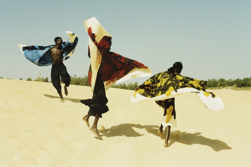Three Black models in flowing garments pose on sand dunes under a clear sky, showcasing fashion and movement in a desert setting.