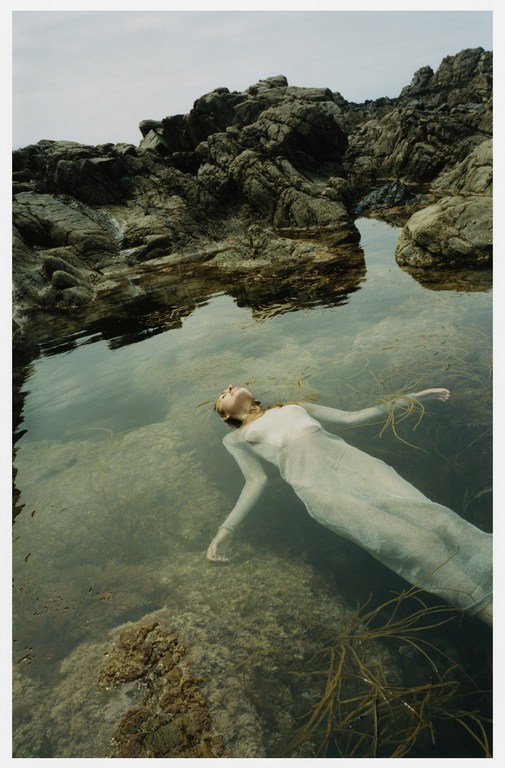 Woman in sheer white dress floats on her back in a shallow pool surrounded by rock formations in a dreamy outdoor setting.