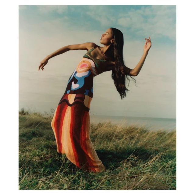Woman in a colorful knit dress poses in a grassy field near the sea on a cloudy day, showcasing a blend of fashion and nature.