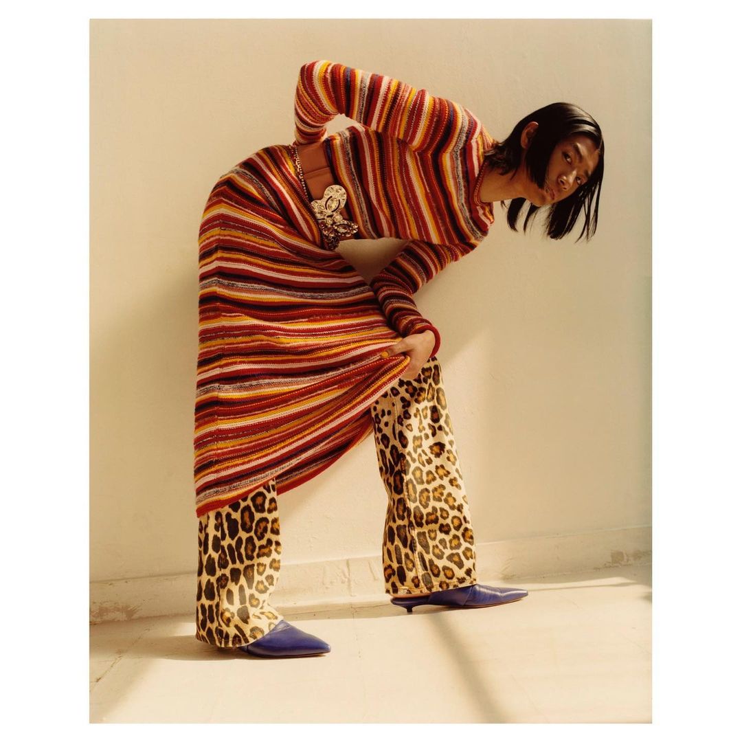 Woman in a striped knit dress and leopard-print pants poses indoors with natural lighting, showcasing a fashion-forward and eclectic style.