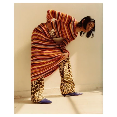 Woman in a striped knit dress and leopard-print pants poses indoors with natural lighting, showcasing a fashion-forward and eclectic style.