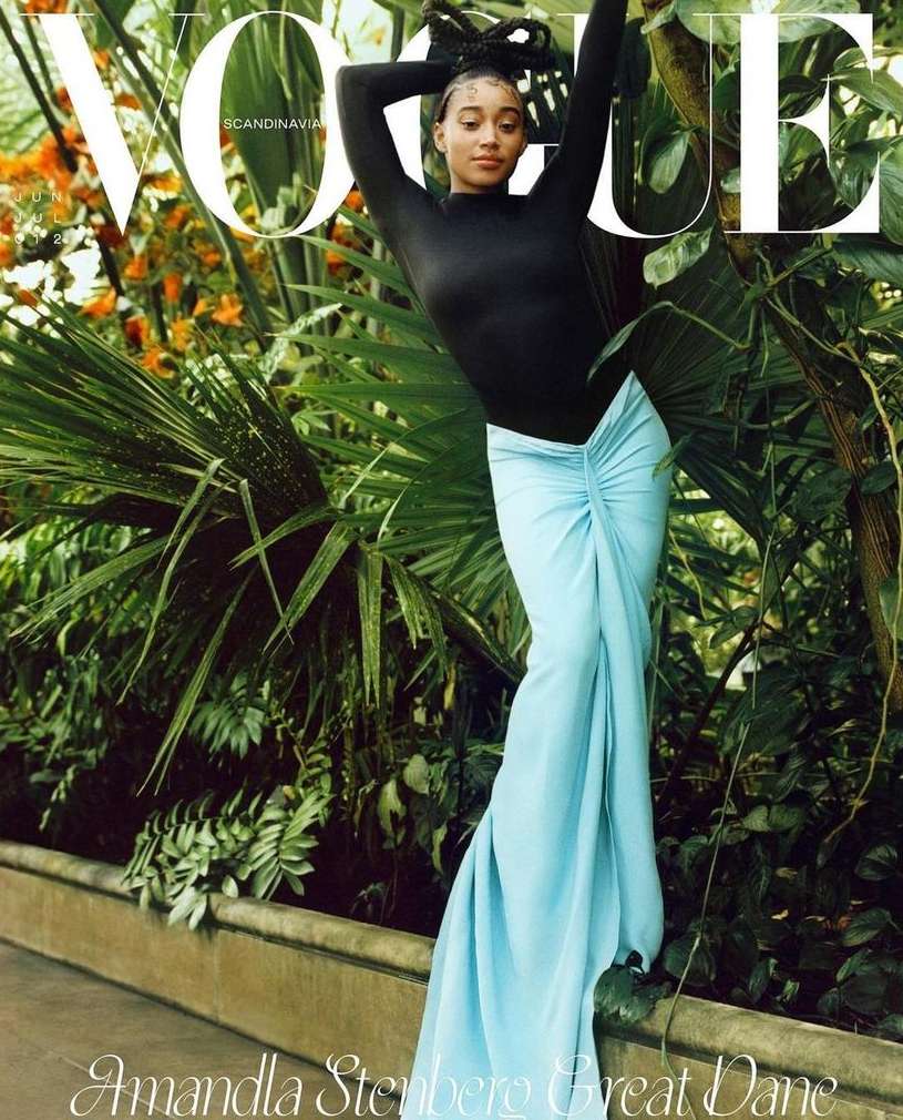 Vogue cover featuring Amandla Stenberg in a black top and draped blue skirt, posing amid lush greenery and foliage.