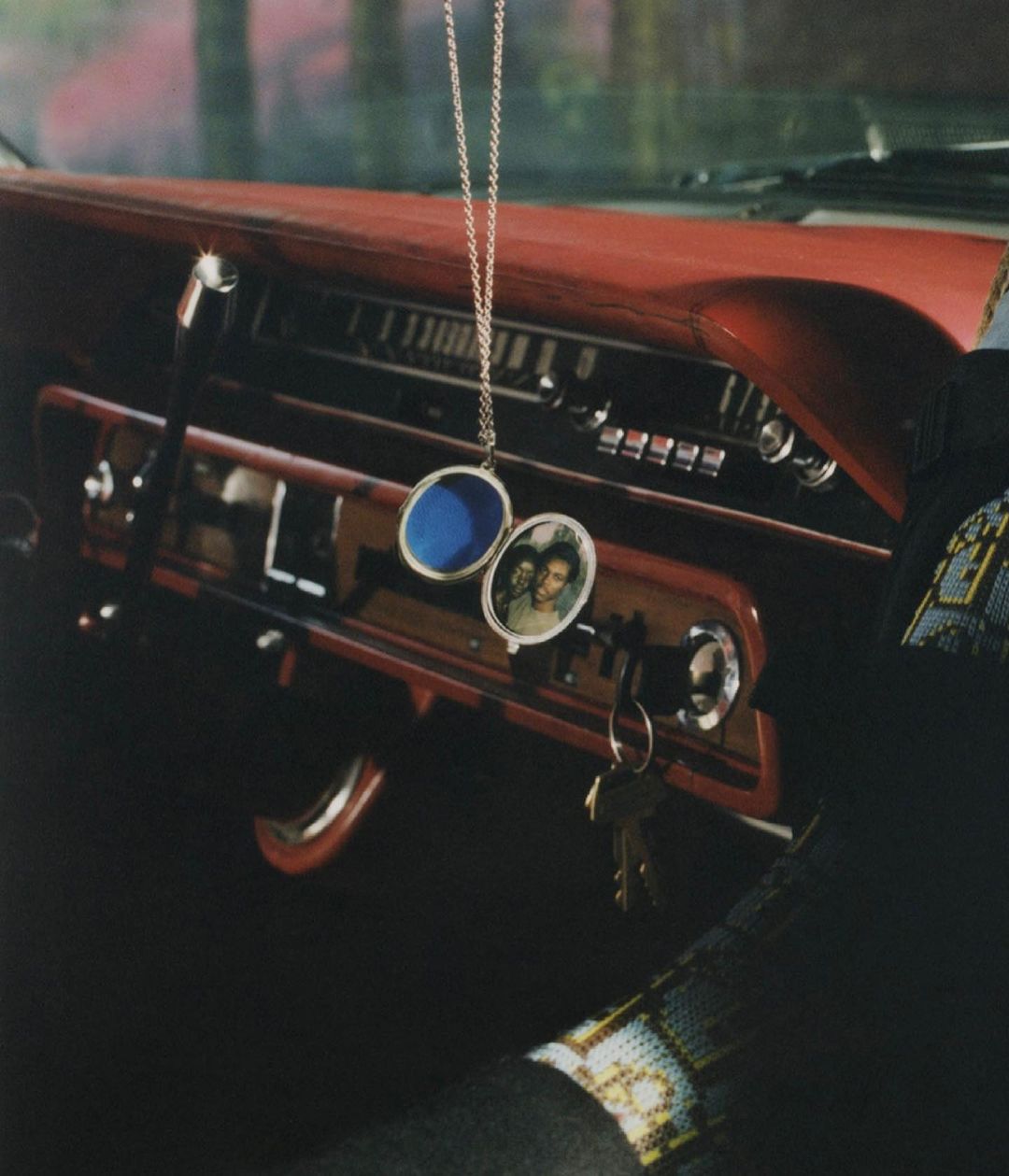 Car interior view showcasing a red dashboard, a dangling photo pendant, keys in the ignition, and a partially visible arm with beaded sleeve.