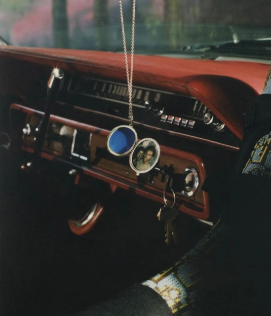 Car interior view showcasing a red dashboard, a dangling photo pendant, keys in the ignition, and a partially visible arm with beaded sleeve.