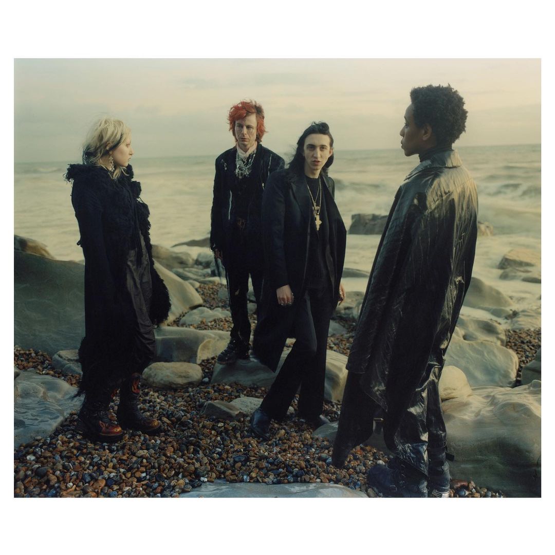 Four people stand on a rocky beach with a muted sky and ocean backdrop. They are dressed in dark clothing, creating a dramatic editorial fashion image.