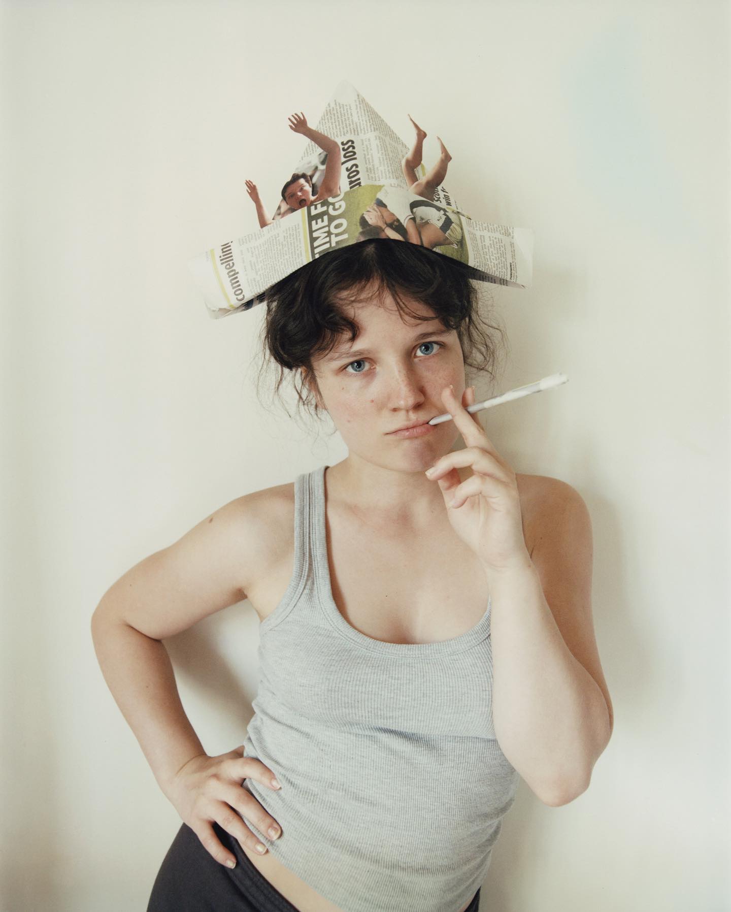 Young adult female poses indoors with newspaper hat, wearing grey tank top and dark shorts, holding a thin white object near lips.