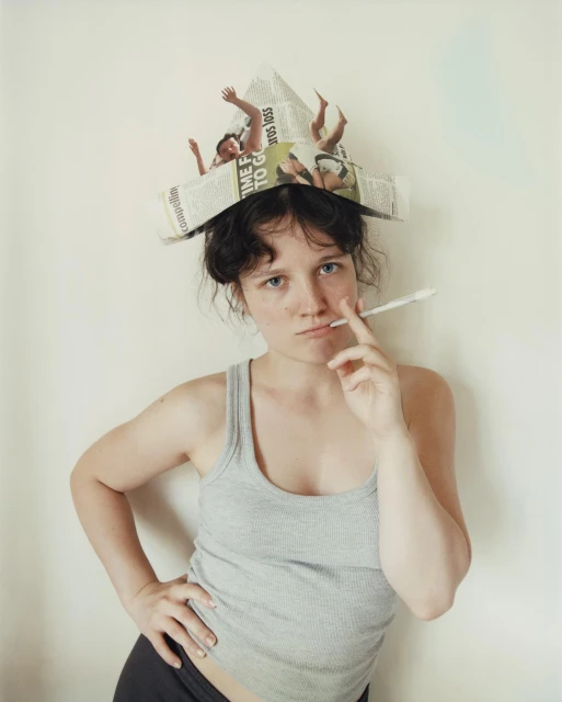 Young adult female poses indoors with newspaper hat, wearing grey tank top and dark shorts, holding a thin white object near lips.