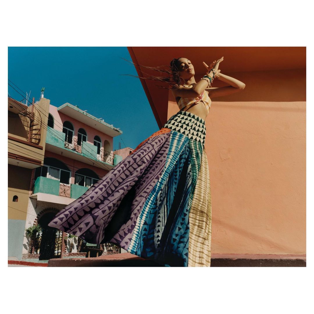 A slender woman in a flowing, multi-colored skirt dances on a vibrant rooftop, set against a backdrop of a peach wall and blue sky.