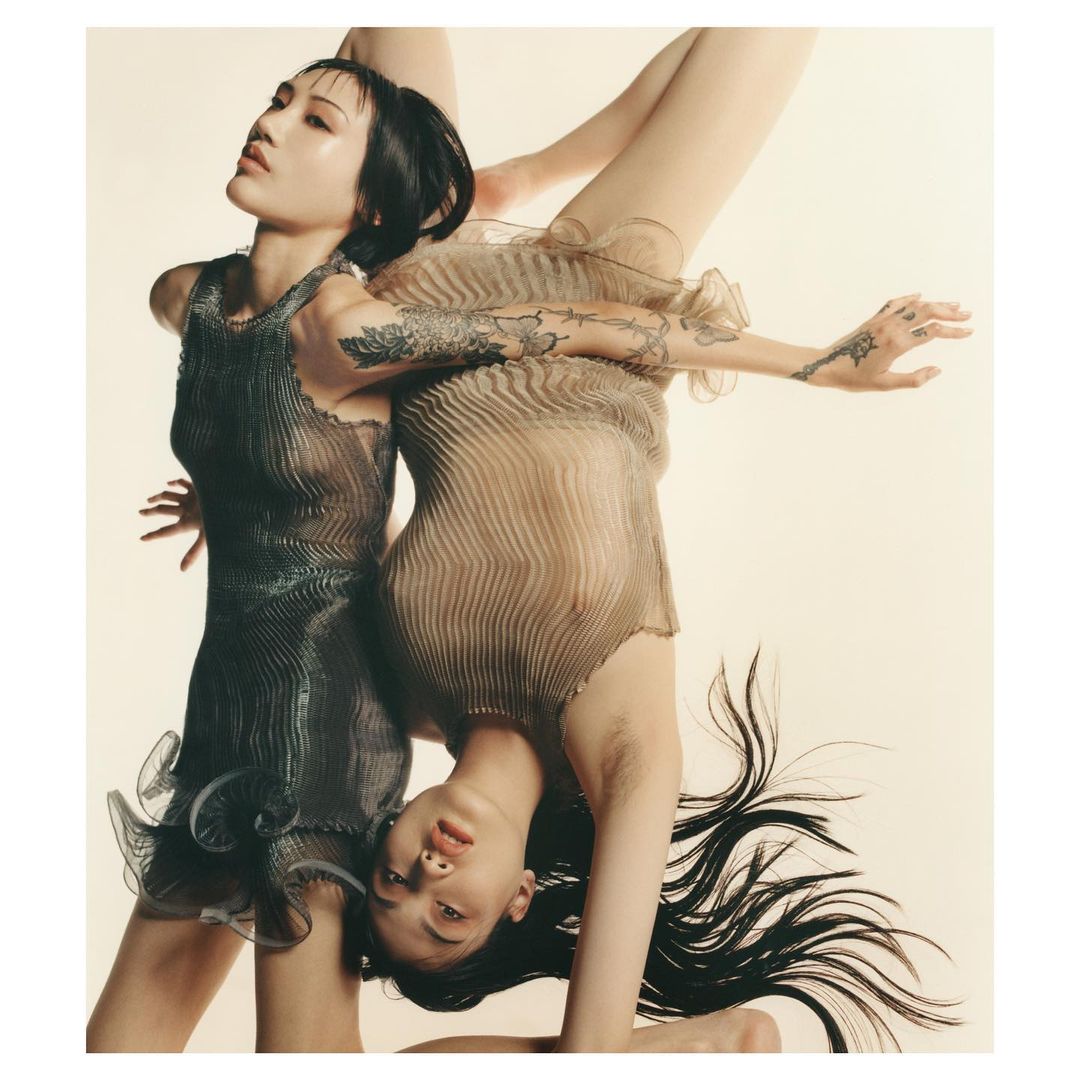 Fashionable shot of two adult women in translucent dresses, one with black hair, one upside down, posing against a neutral-toned backdrop.