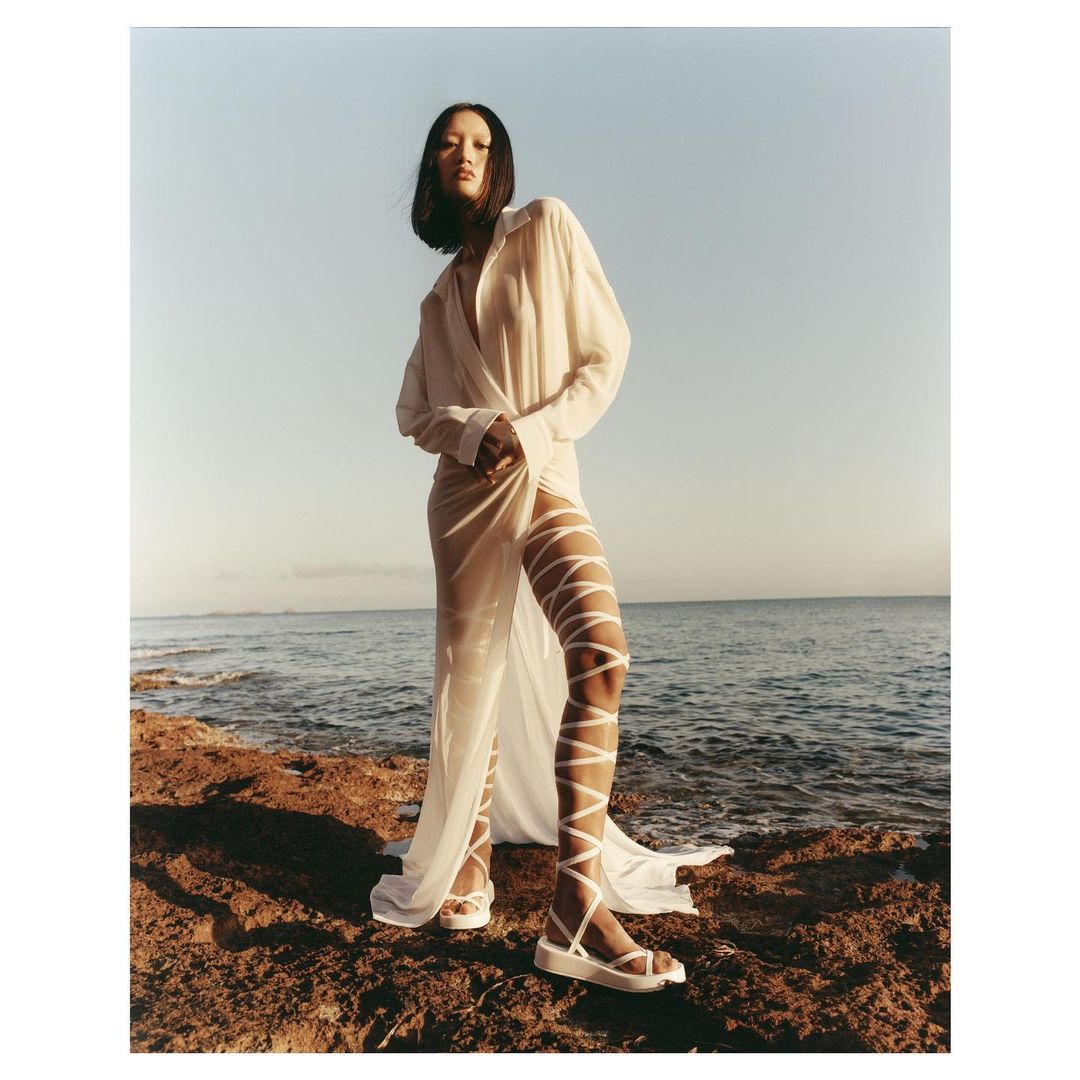 A woman in a white robe and sandals stands on a rocky shore, posing against a blue sea and sky in natural light.
