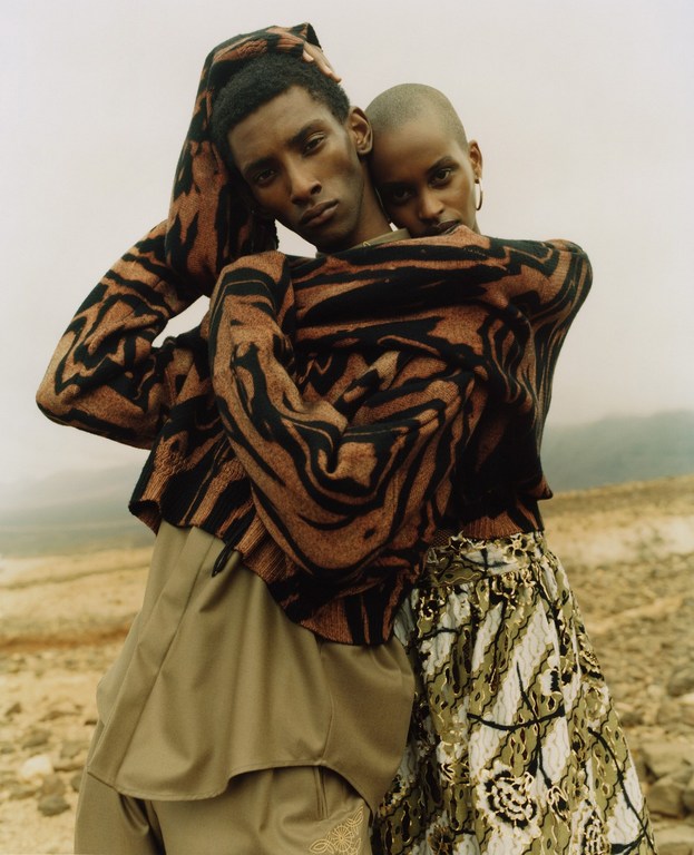A stylish couple in a desert landscape, showcasing patterned sweaters and skirts, with warm, earthy tones and a minimalist aesthetic.
