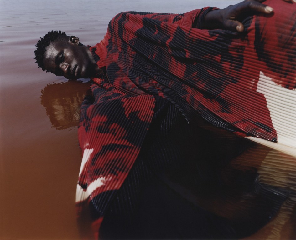 A Black male fashion model is lying in shallow water in a red and black patterned top, with a subtle reflection visible.