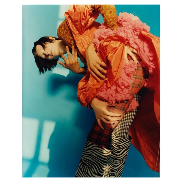 Fashionable person with dark hair poses with hands in frame, wearing an orange jacket with pink ruffles and patterned pants on a blue background.