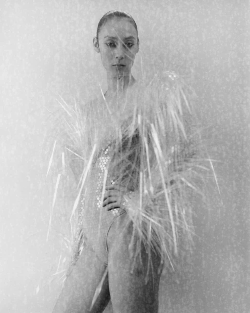 Black-and-white portrait of a woman in a sparkly leotard, posing with hand on hip, partially obscured by decorative grass.