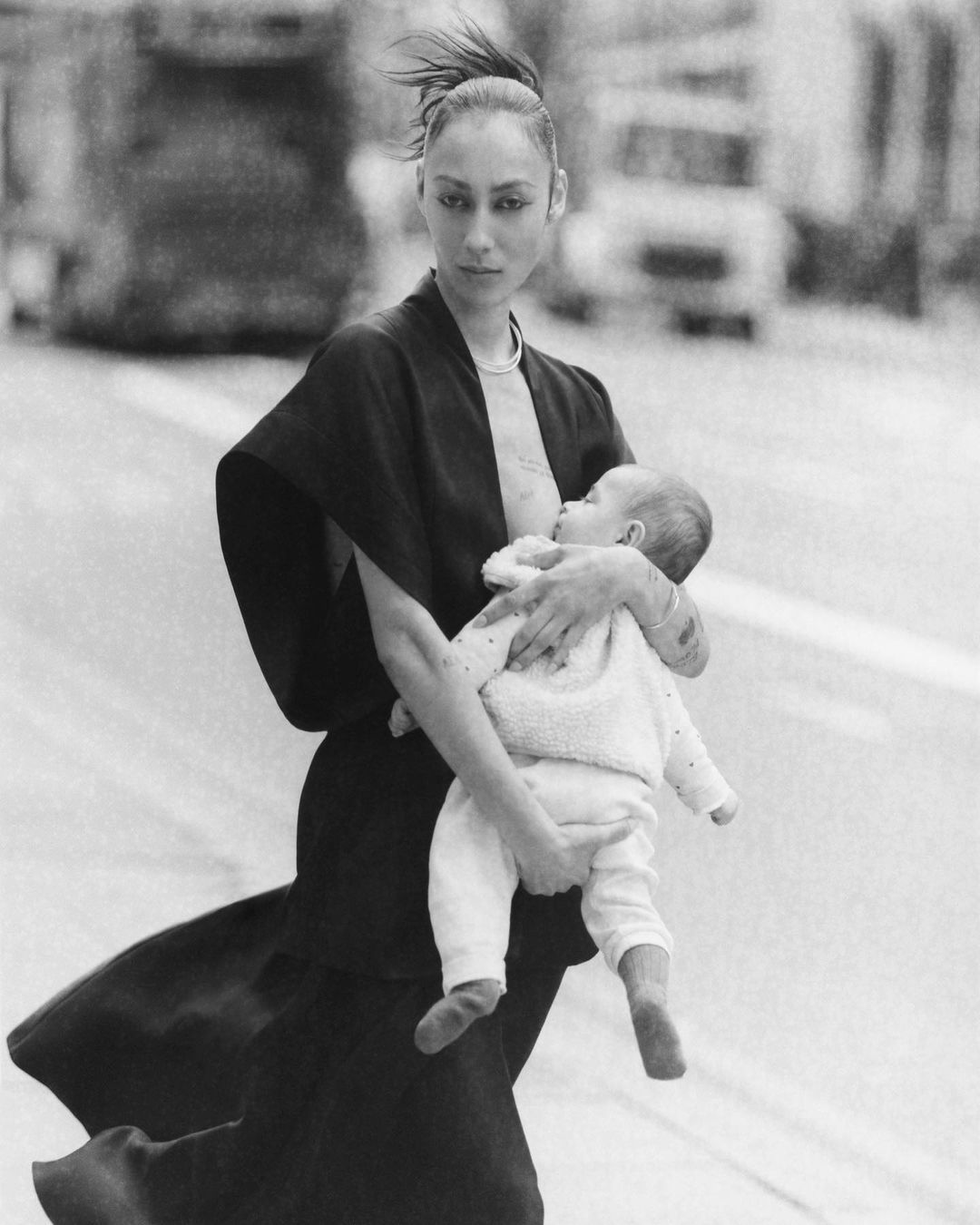 Black and white shot of woman outdoors breastfeeding her baby while walking across a street, with blurred vehicles in background.