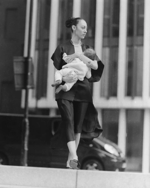 Black and white photo of a stylish adult woman holding a baby while walking on a city street, with an automobile in the background.