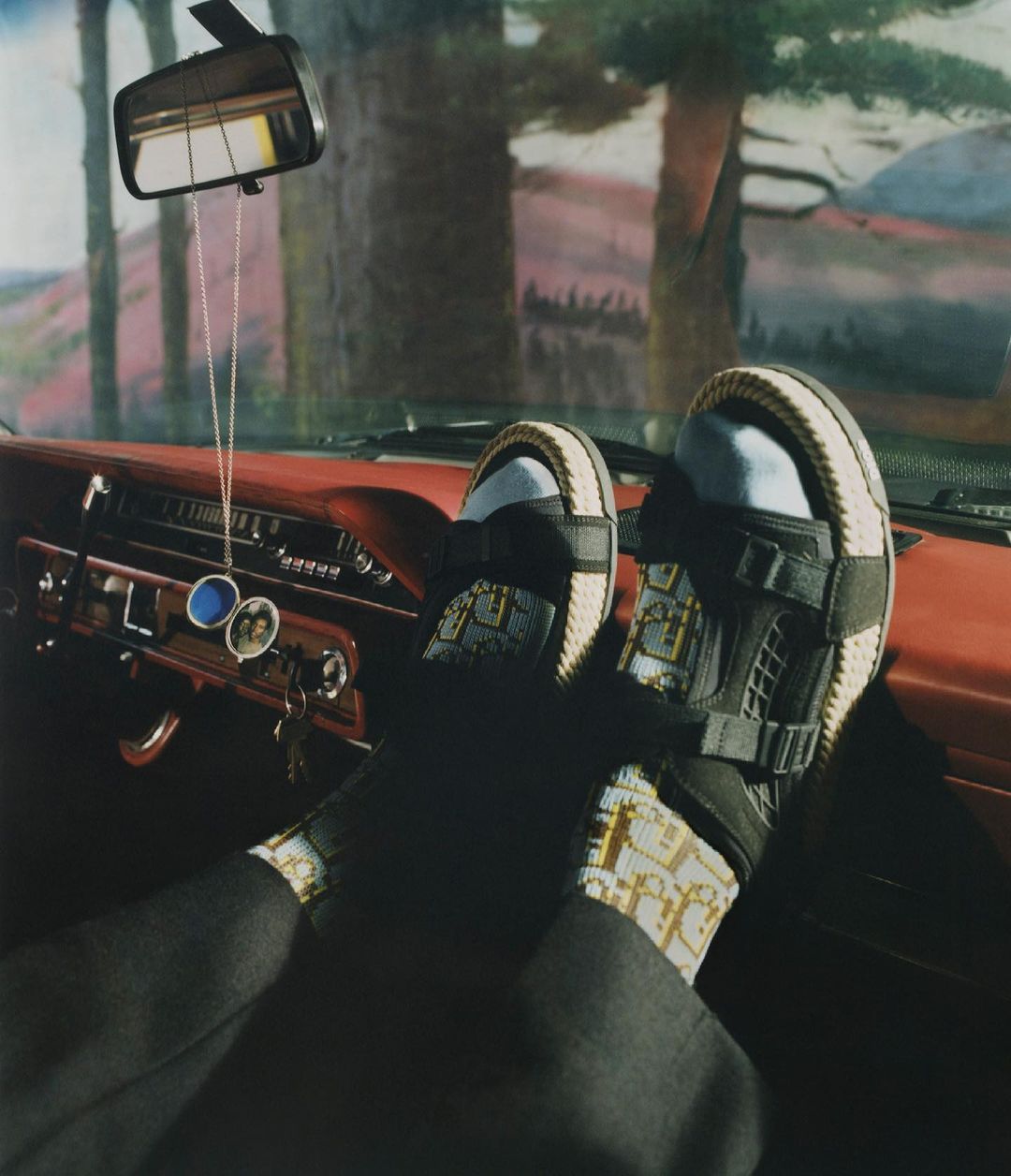 Person resting feet in luxury sandals on the dashboard of a vintage car, with view through the windshield to a background landscape.