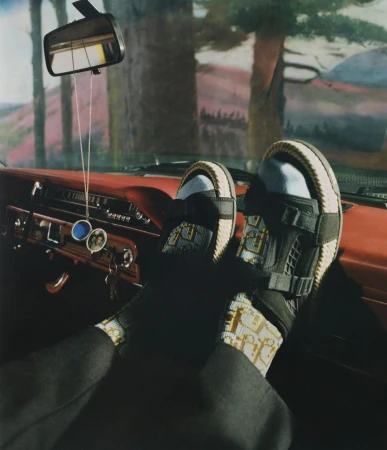Person resting feet in luxury sandals on the dashboard of a vintage car, with view through the windshield to a background landscape.