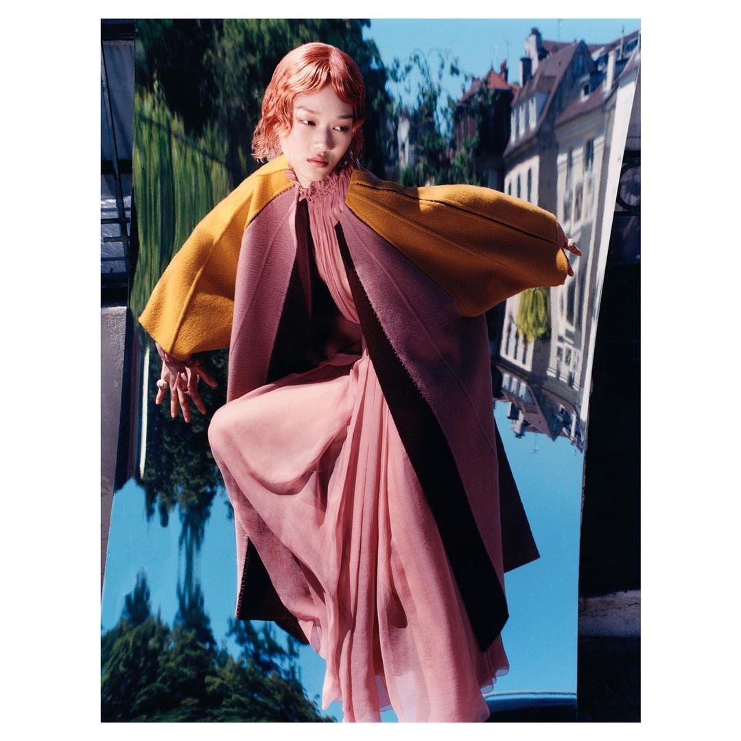 Woman with short red hair poses in flowing rose-pink dress with yellow and rose color-block cloak, against backdrop of blurred buildings.