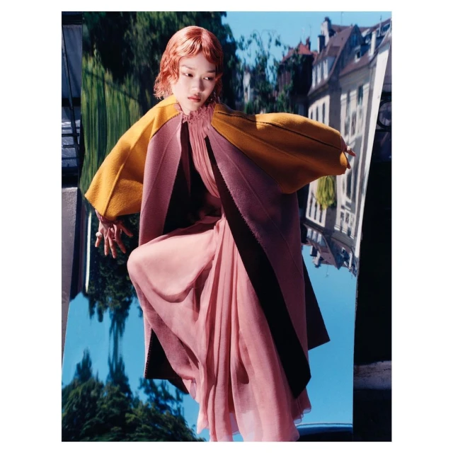 Woman with short red hair poses in flowing rose-pink dress with yellow and rose color-block cloak, against backdrop of blurred buildings.