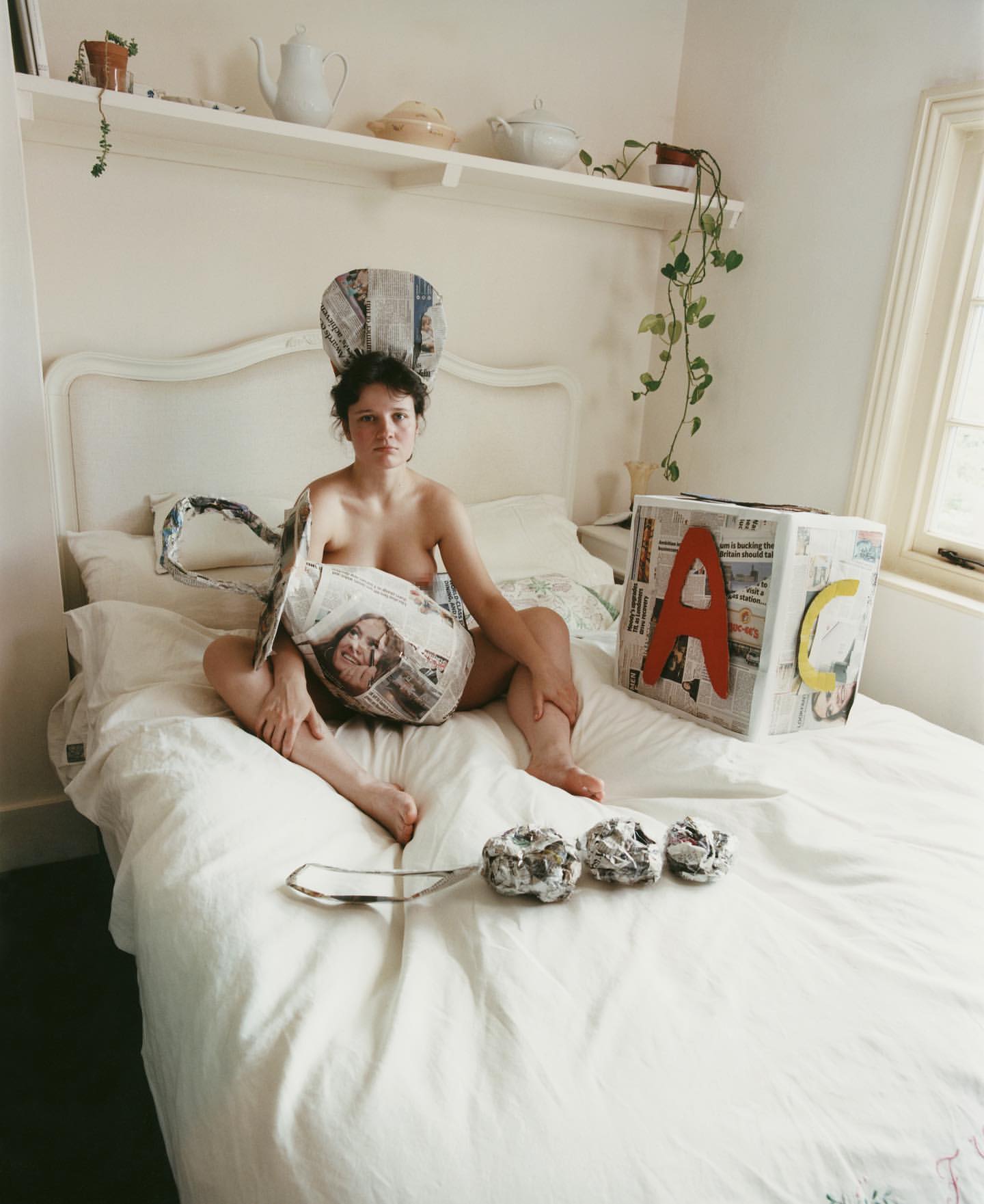 Woman sits on bed adorned with newspaper objects including a hat, bag, and building block with a shelf and window in the background.