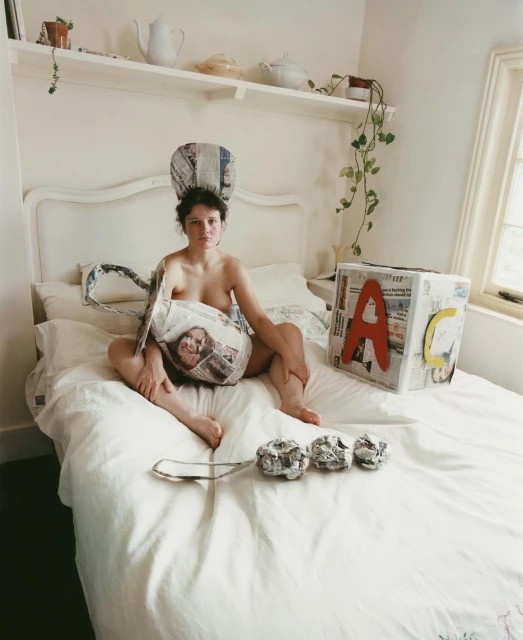 Woman sits on bed adorned with newspaper objects including a hat, bag, and building block with a shelf and window in the background.