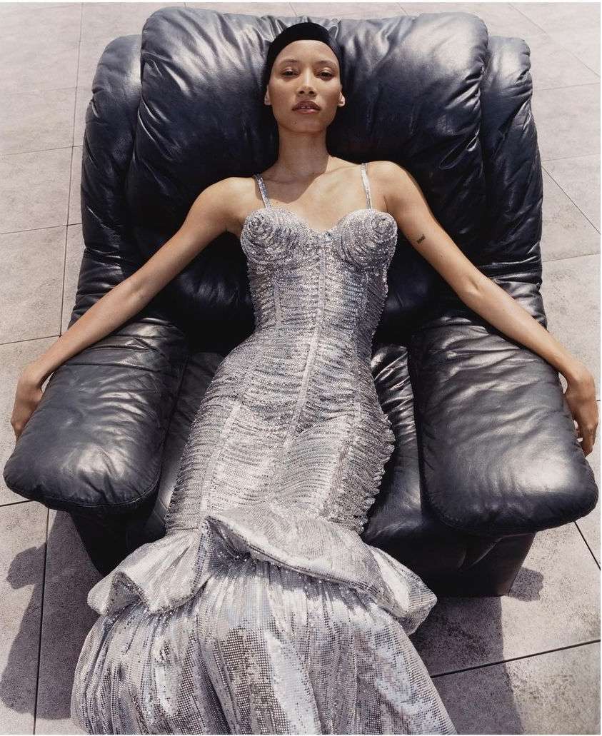 Elegant woman in a shimmering silver dress reclines in a black leather chair, highlighted by natural light and set against a tiled floor.