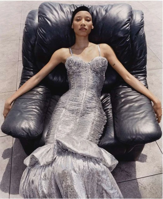 Elegant woman in a shimmering silver dress reclines in a black leather chair, highlighted by natural light and set against a tiled floor.