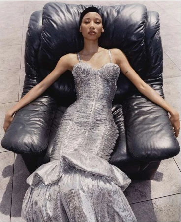 Elegant woman in a shimmering silver dress reclines in a black leather chair, highlighted by natural light and set against a tiled floor.