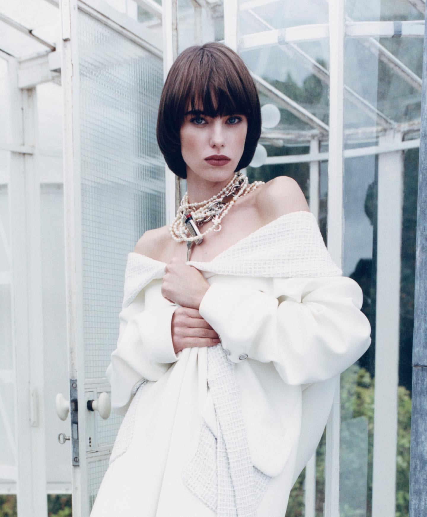 Portrait of an adult female fashion model with short brown hair wearing an off-shoulder white coat and necklace, posing by a glass structure.
