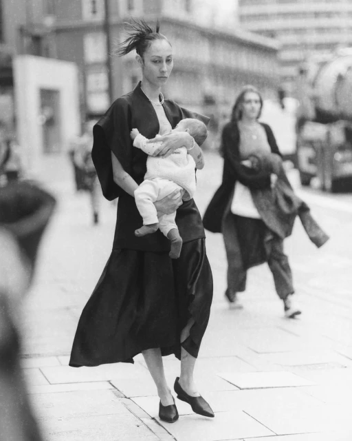 Woman in black dress holds baby in a street setting with blurred figures in background, creating a moment captured in black and white.