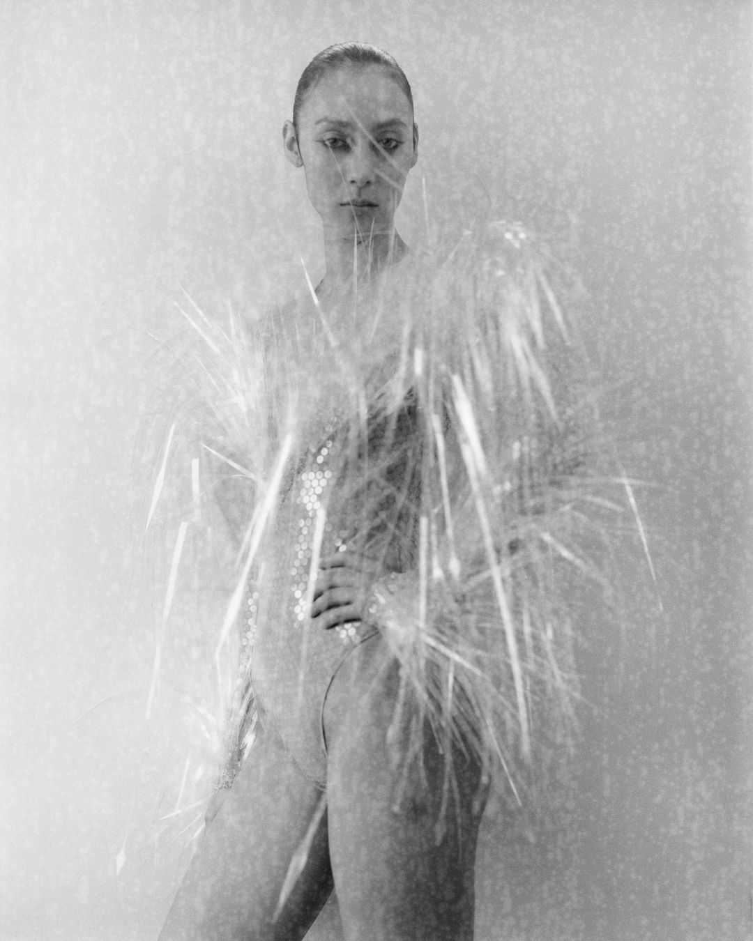 Black and white portrait of a woman in a sequin bodysuit, partially obscured by decorative strands, against a neutral background.