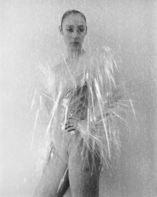 Black and white portrait of a woman in a sequin bodysuit, partially obscured by decorative strands, against a neutral background.