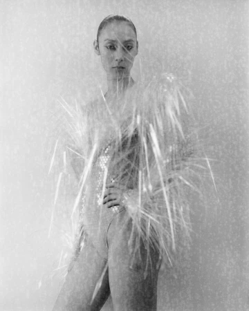 Black and white portrait of a woman in a sequin bodysuit, partially obscured by decorative strands, against a neutral background.