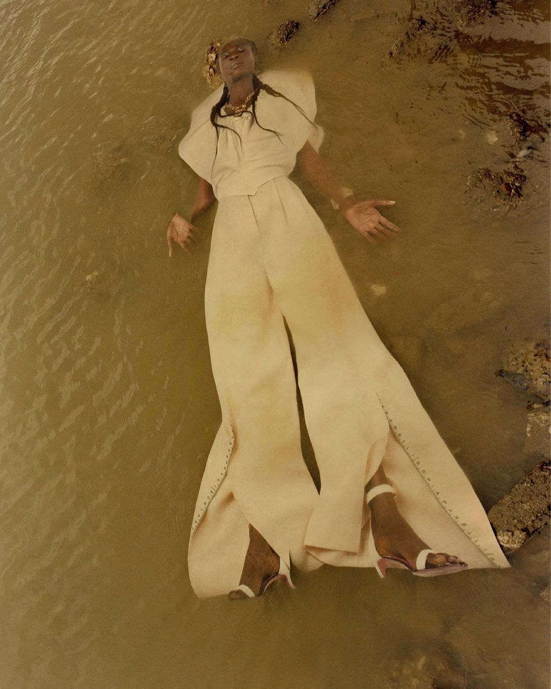 Fashion shot: Woman in flowing white pants and top lying in shallow water with rocky shore. High-angle shot. Editorial fashion.