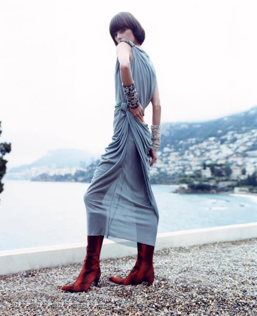 Fashion shot of an adult woman with short dark hair wearing a grey draped dress and reddish brown boots standing on a stone surface.
