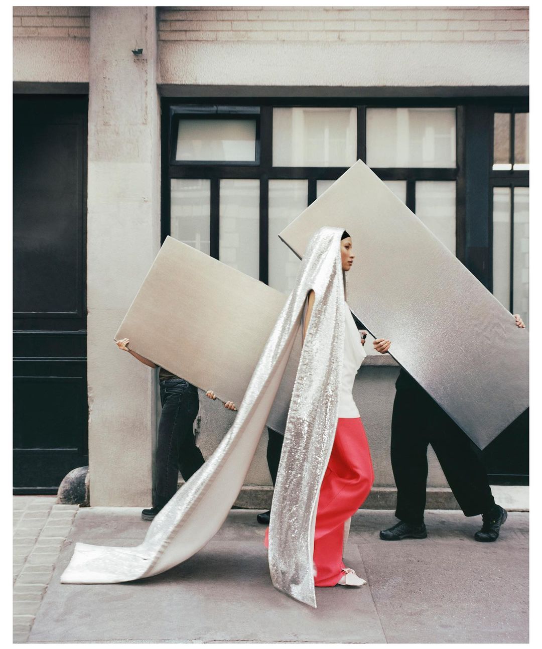 Editorial fashion shot of a woman in a white top and coral pants walking down a city street with two people carrying panels.