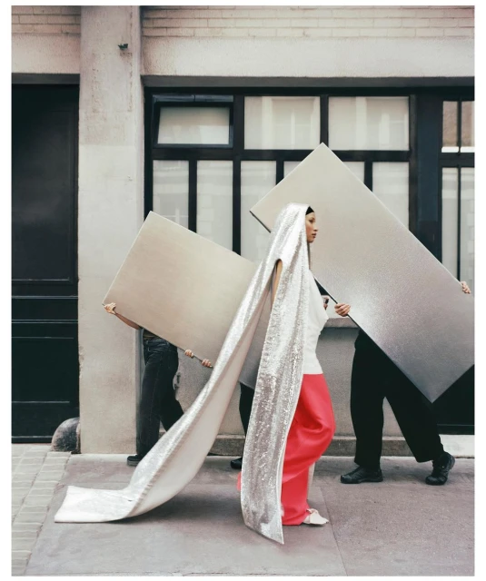 Editorial fashion shot of a woman in a white top and coral pants walking down a city street with two people carrying panels.