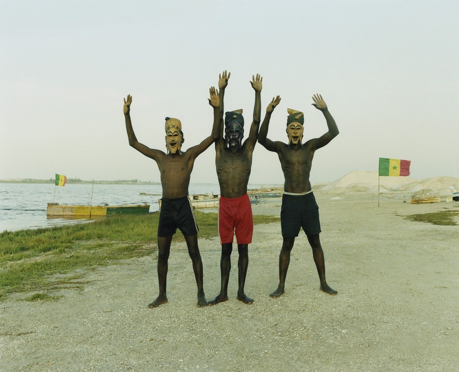 Three men with face paint stand on a beach with arms raised near a lake and flags; landscape features a grassy shoreline and sandy ground.