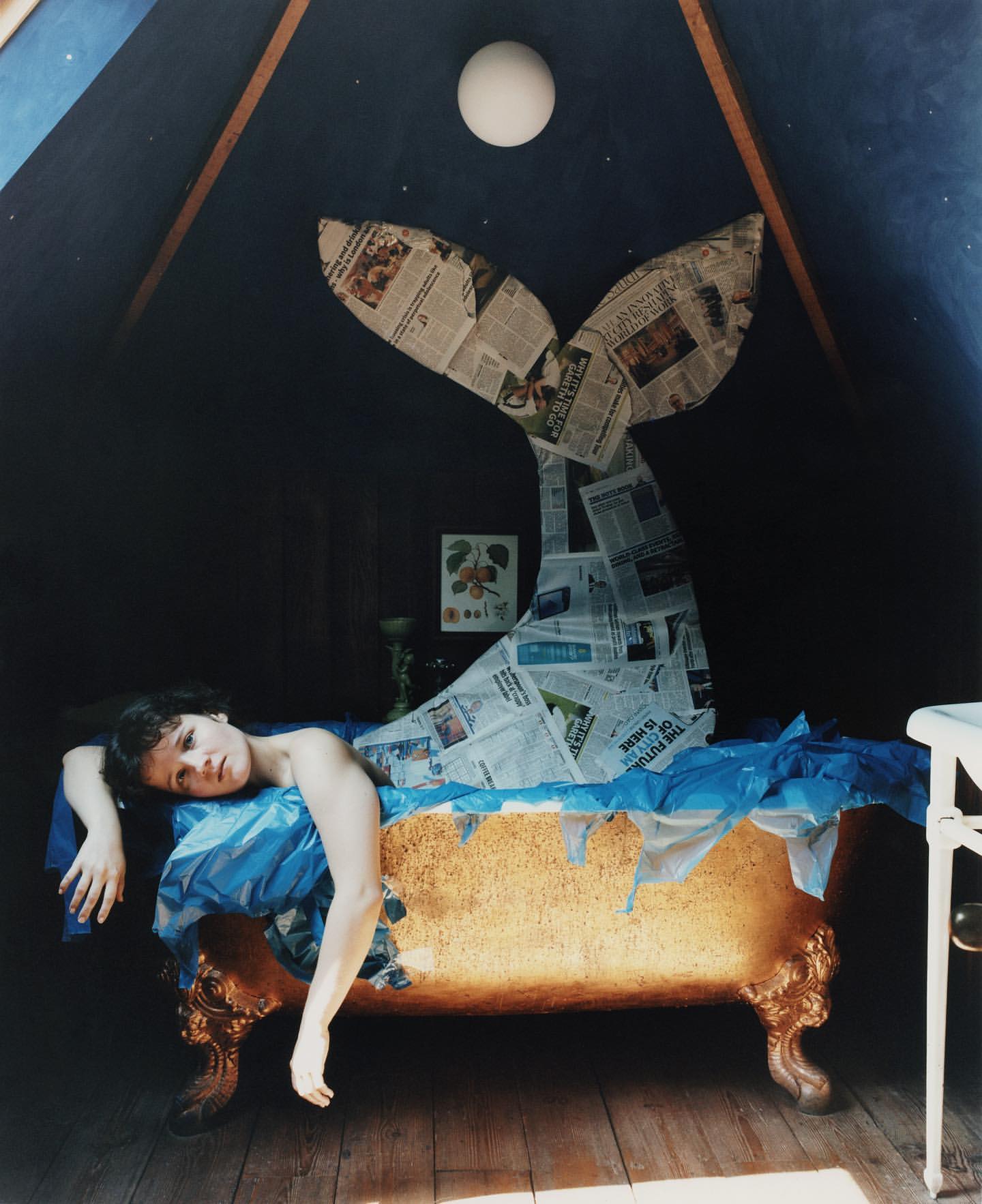An unusual photograph depicts a person reclining in a bathtub beneath a whale tail sculpture made of newspaper inside a dark room.